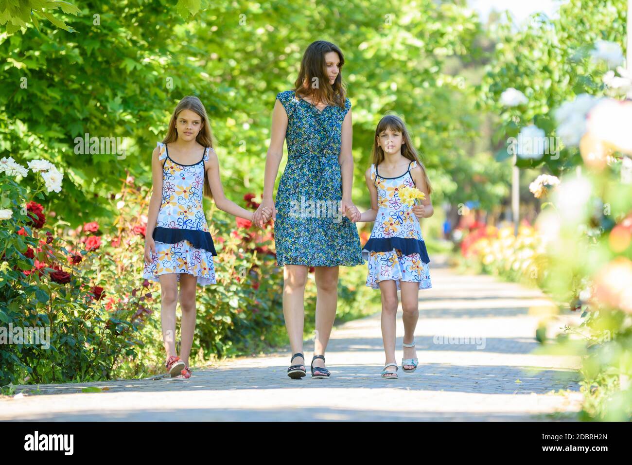 young woman walks down the path in the flowering garden by the hand with her two daughters Stock Photo