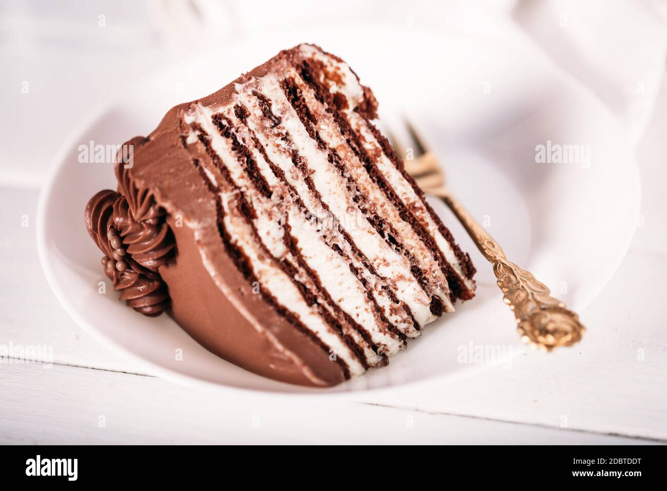 A piece of chocolate cake on a white plate with a gold fork on a white ...