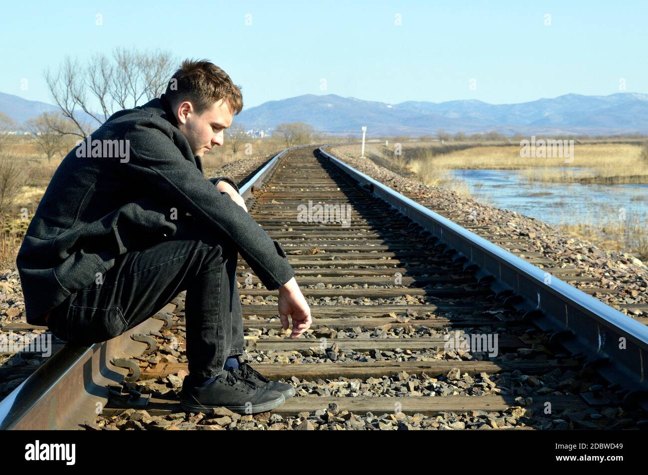Young man in coat looking down with upset face,sitting on rails. Depressive concept loss of money, loneliness, unemployment. Copy space Stock Photo