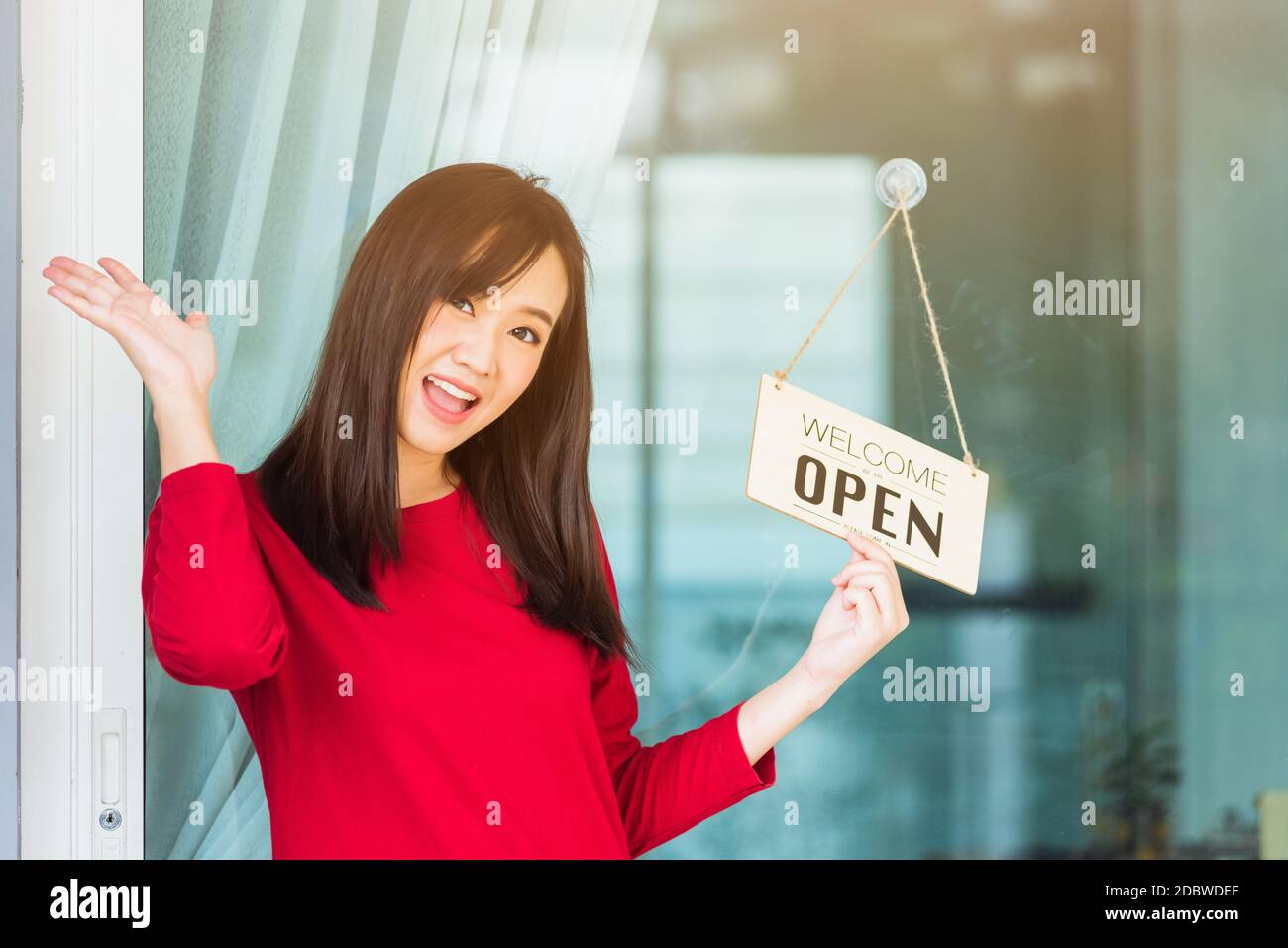 Asian young woman glad and smiling she notice sign wood board label 'WELCOME OPEN' hanging through glass door front shop, Business turning open after Stock Photo