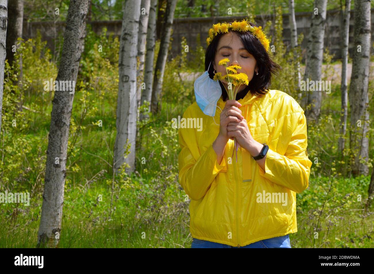 Brunette woman with closed eyes,in yellow jacket with wreath of dandelions and removed medical mask smelling bouquet of  dandelions. coronavirus pande Stock Photo