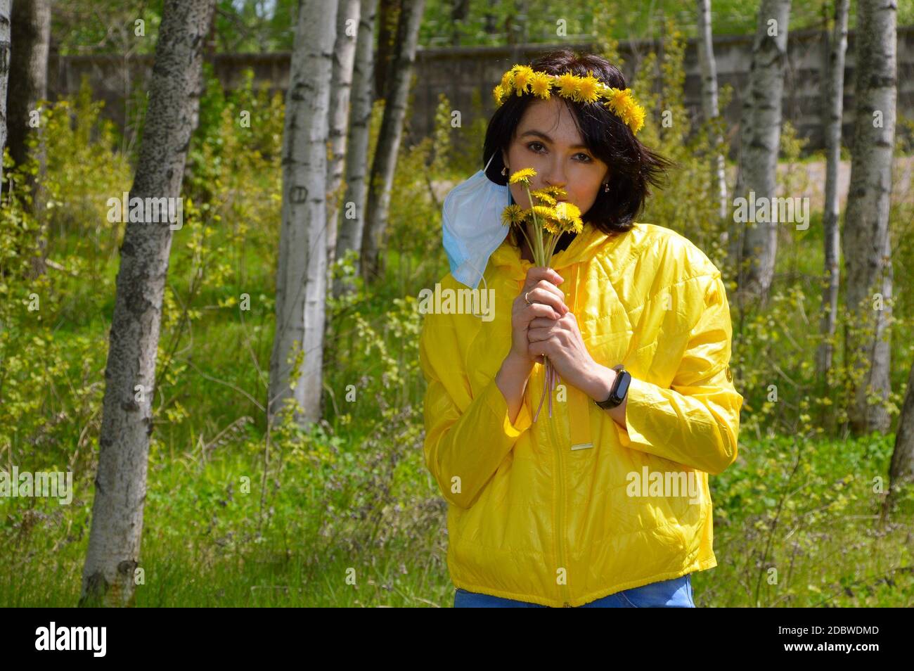 Brunette woman with closed eyes,in yellow jacket with wreath of dandelions and removed medical mask smelling bouquet of  dandelions. coronavirus pande Stock Photo
