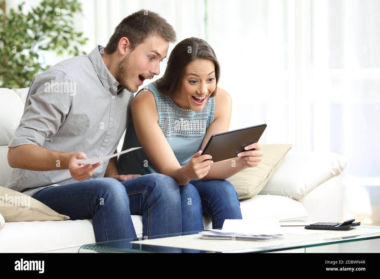 Excited couple checking tablet and receipts sitting in the sofa in the  living room at home Stock Photo - Alamy