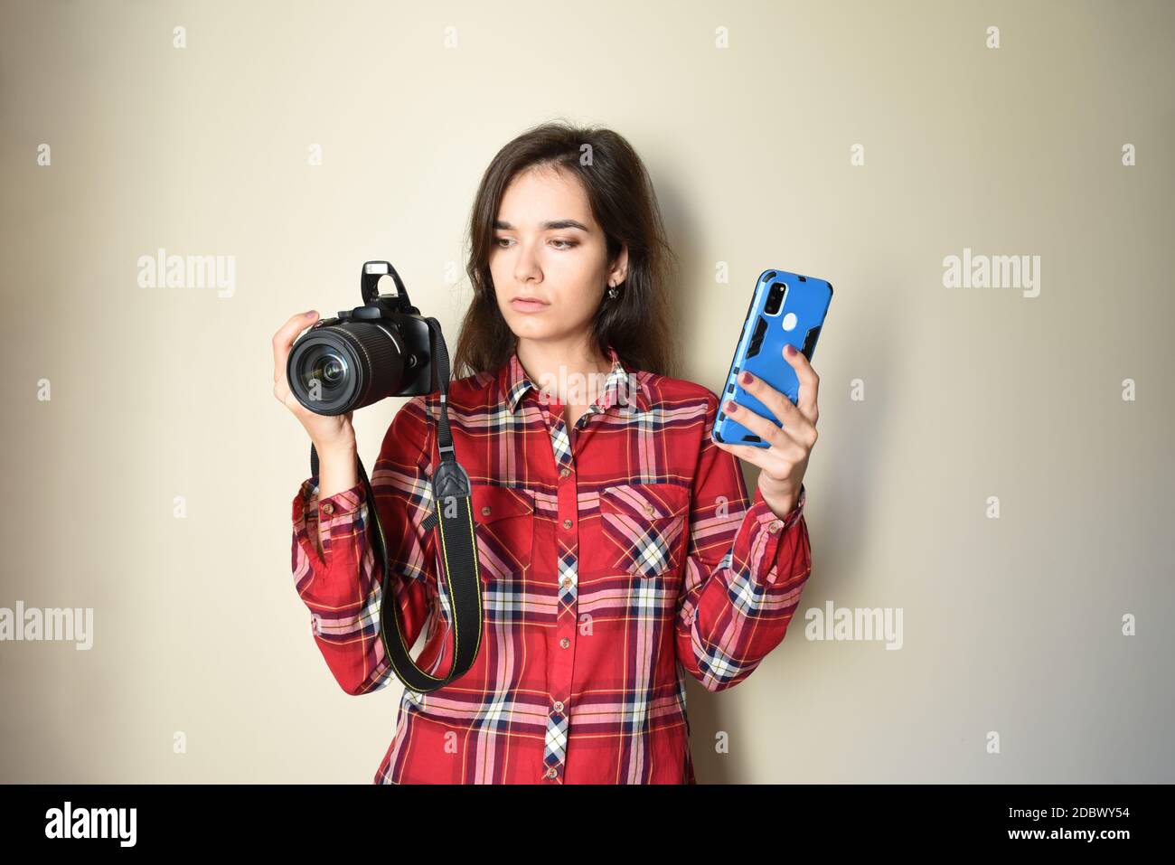 Young brunette woman photographer in red shirt choosing between camera and mobile phone on beige background. Concept of choice and difference Stock Photo