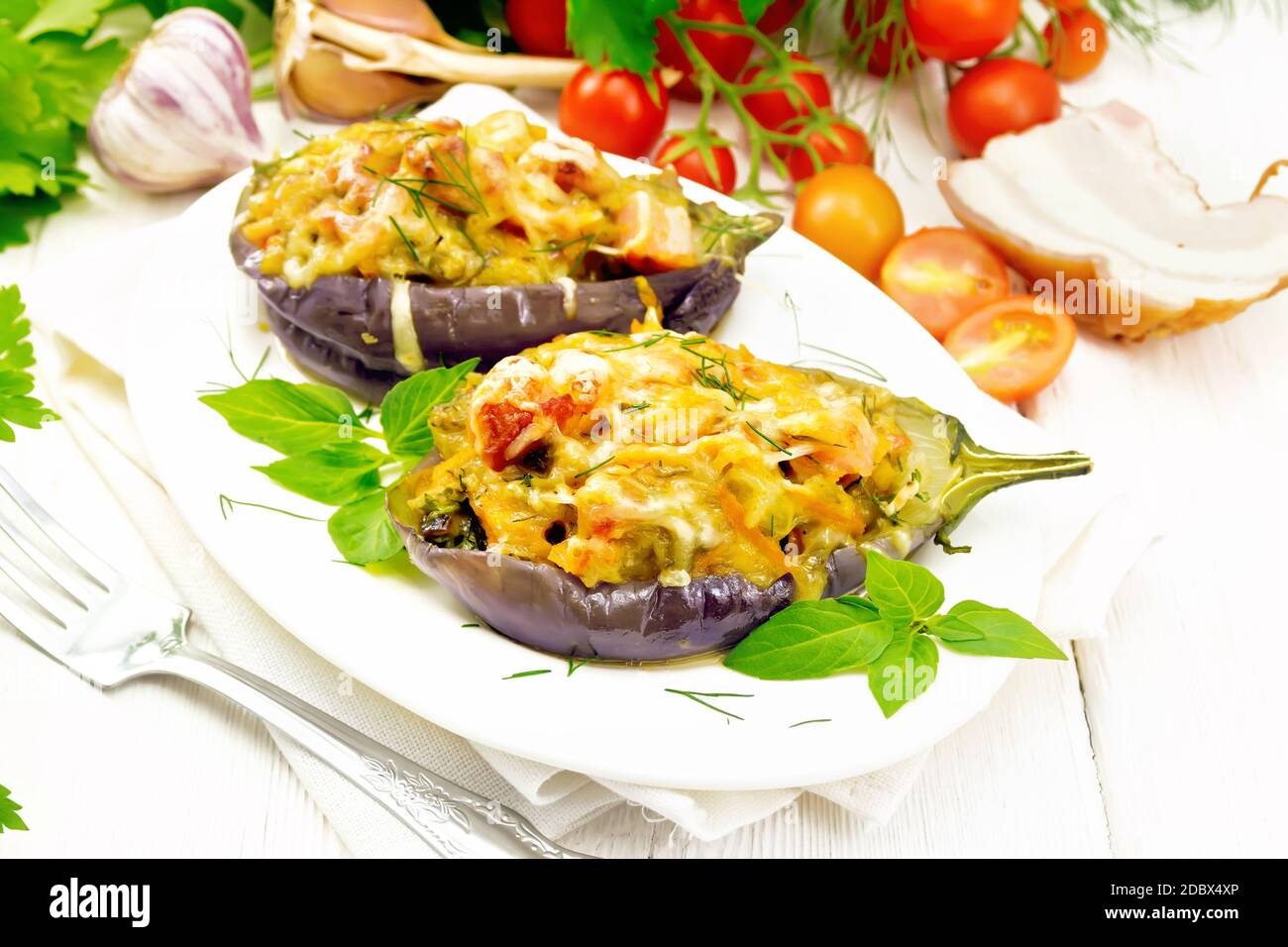 Stuffed eggplant with smoked bacon, tomatoes, onions, carrots with garlic, cheese and herbs in an oval plate on a napkin on wooden board background Stock Photo