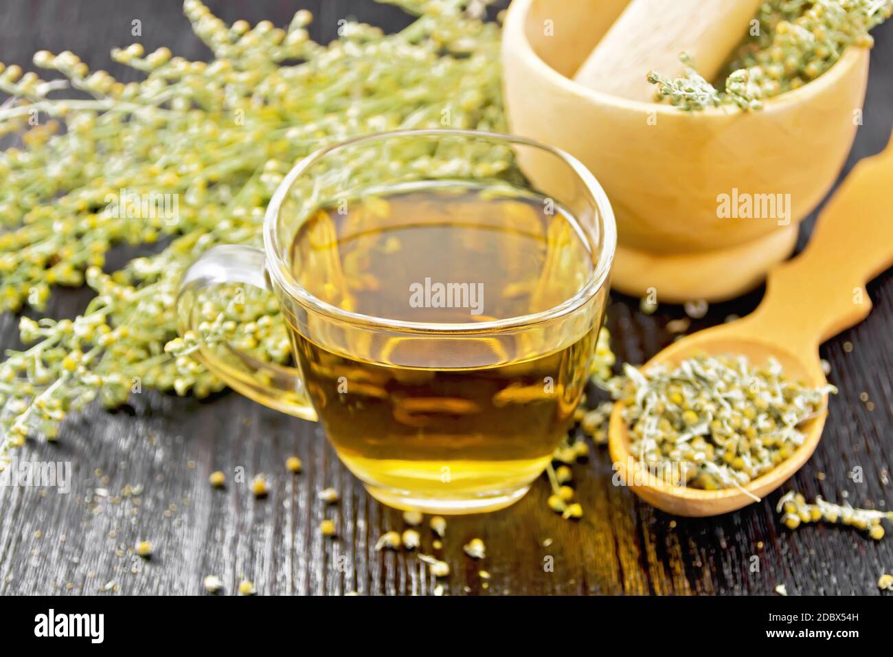 Gray wormwood herbal tea in a glass cup, dry sagebrush flowers in spoon, fresh flowers in a mortar and on table against dark wooden board Stock Photo