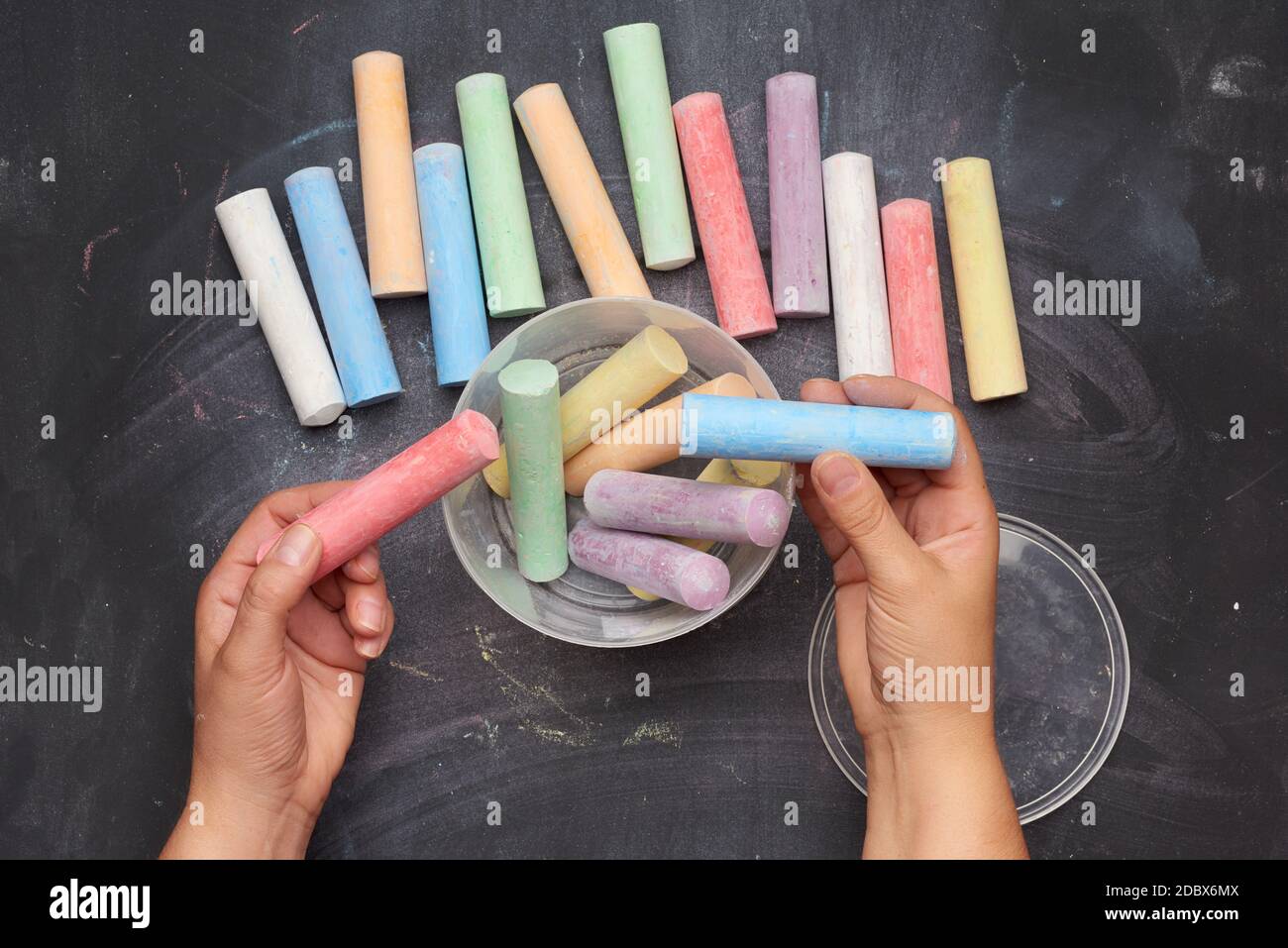 multicolored chalk in a plastic bucket on a black background, female hands fold objects into a container, top view Stock Photo