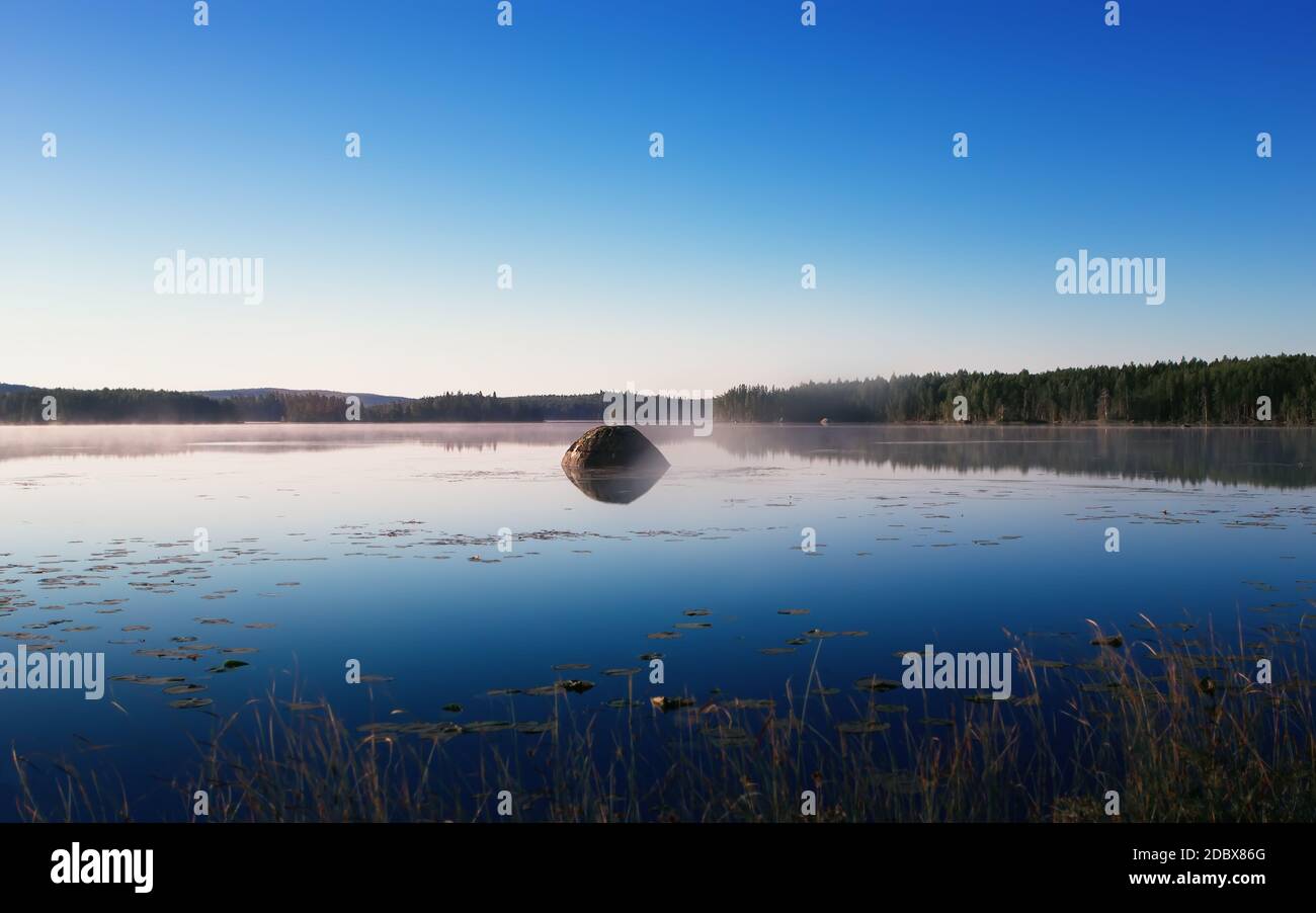 Cool misty summer morning reflected in the northern forest lake. Large stone boulder island in the mirror water under a clear blue sky. Natural backgr Stock Photo