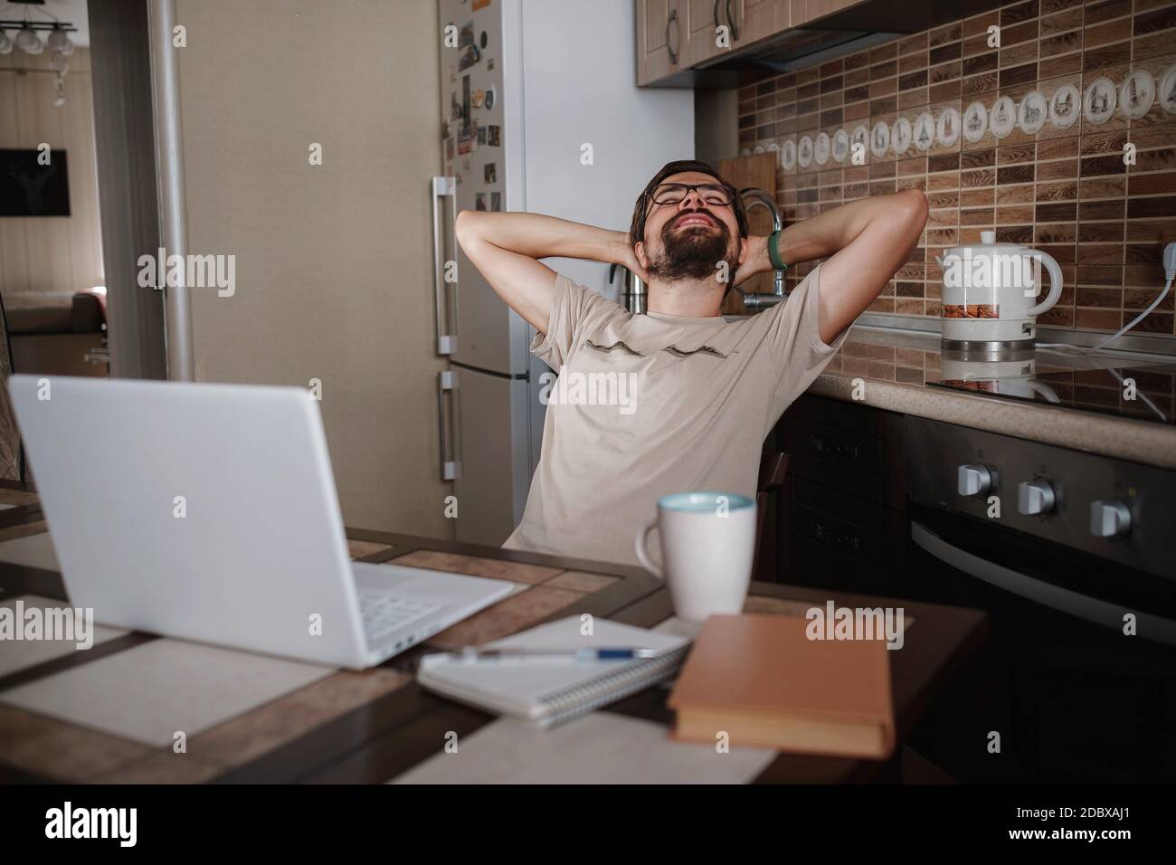 Smiling hipster freelancer holding hands behind head sitting at office desk behind laptop. Happy employee feeling no stress, relaxing, watching funny Stock Photo