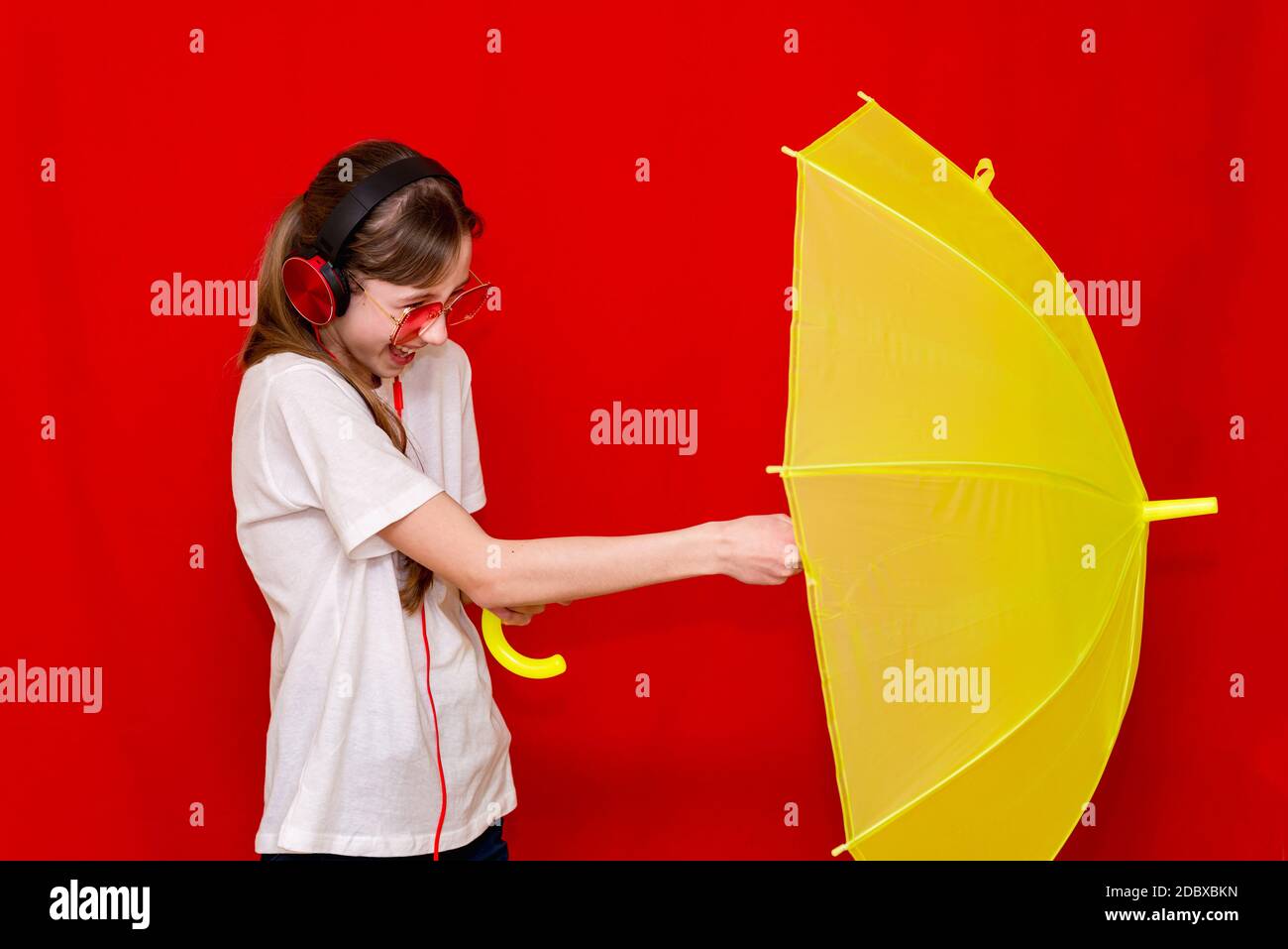 Teenage girl listens to music in headphones with umbrella. red studio background wearing white t shirt.studio shot. Stock Photo