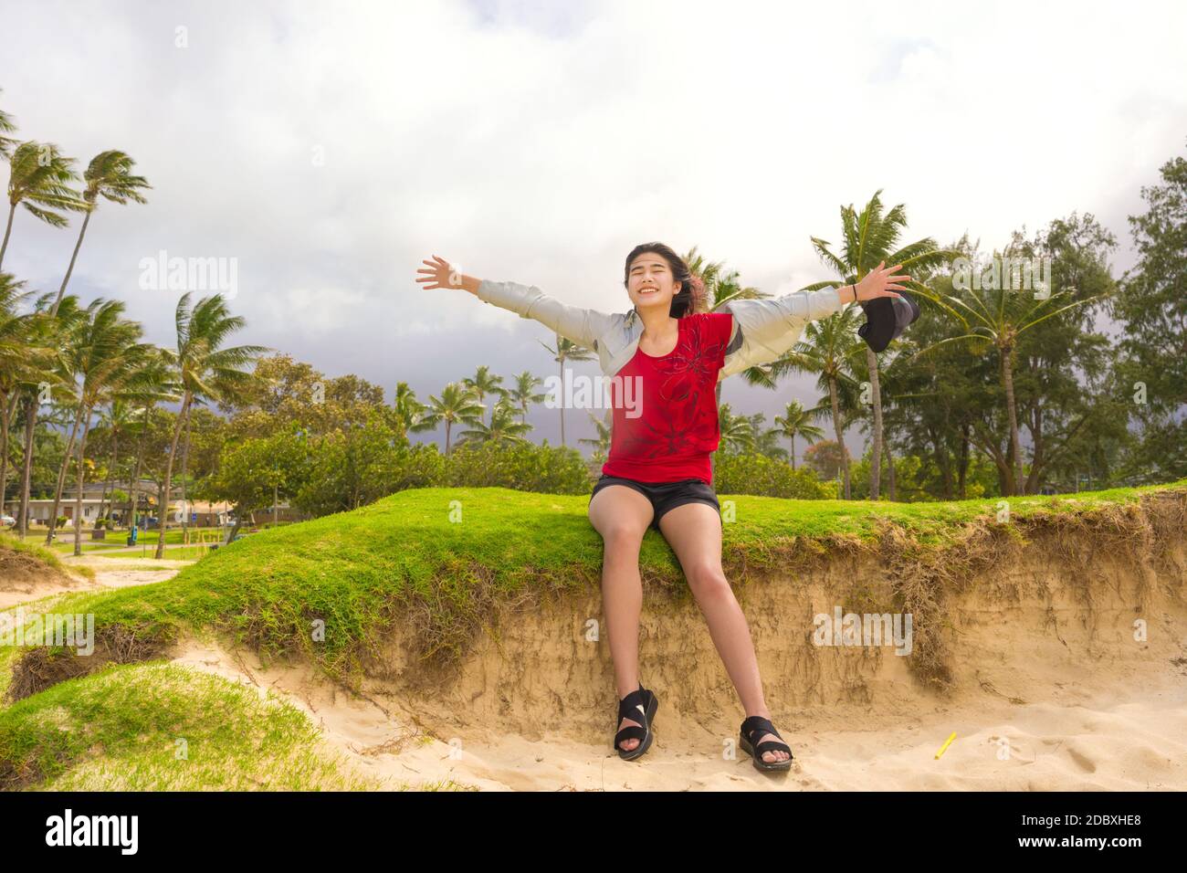 Biracial teen girl sitting along grassy dunes on sunny Hawaiian beach by the ocean, arms raised enjoying the wind Stock Photo