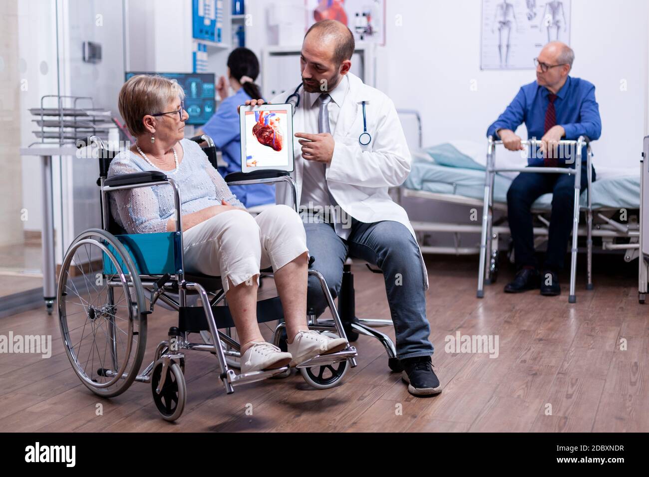 Coronary artery disease explanation from doctor to disabled senior woman in wheelchair during medical examination. Man with disabilities ,walking frame sitting in hospital bed. Health care system. Stock Photo