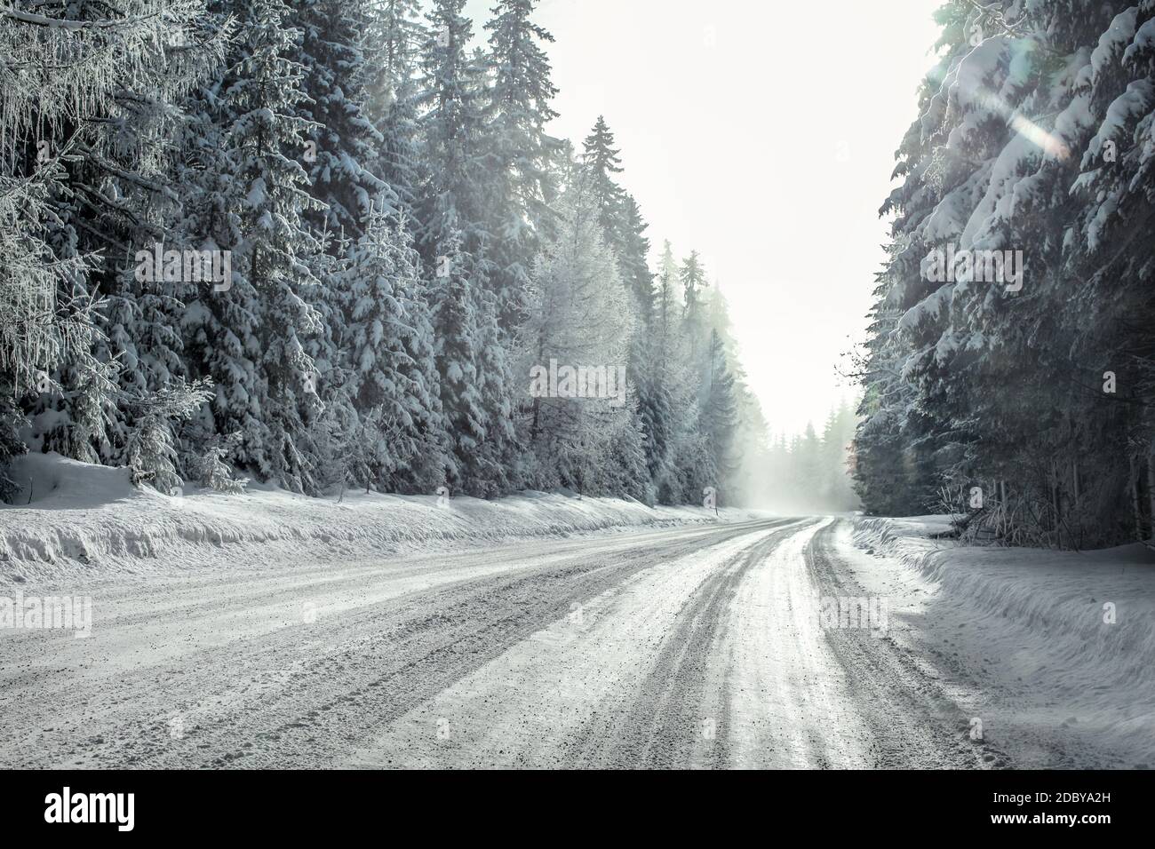 View from a car riding through snow covered winter road curve, lit by strong sun backlight and fog and haze in distance - dangerous driving conditions Stock Photo