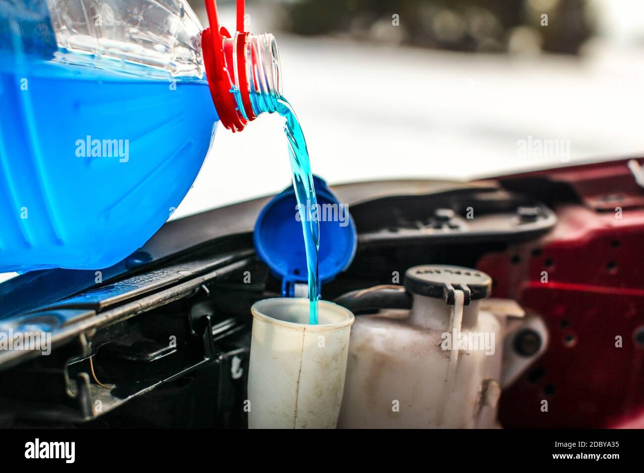 Detail on pouring antifreeze liquid screen wash into dirty car from blue and red anti freeze water container. Stock Photo