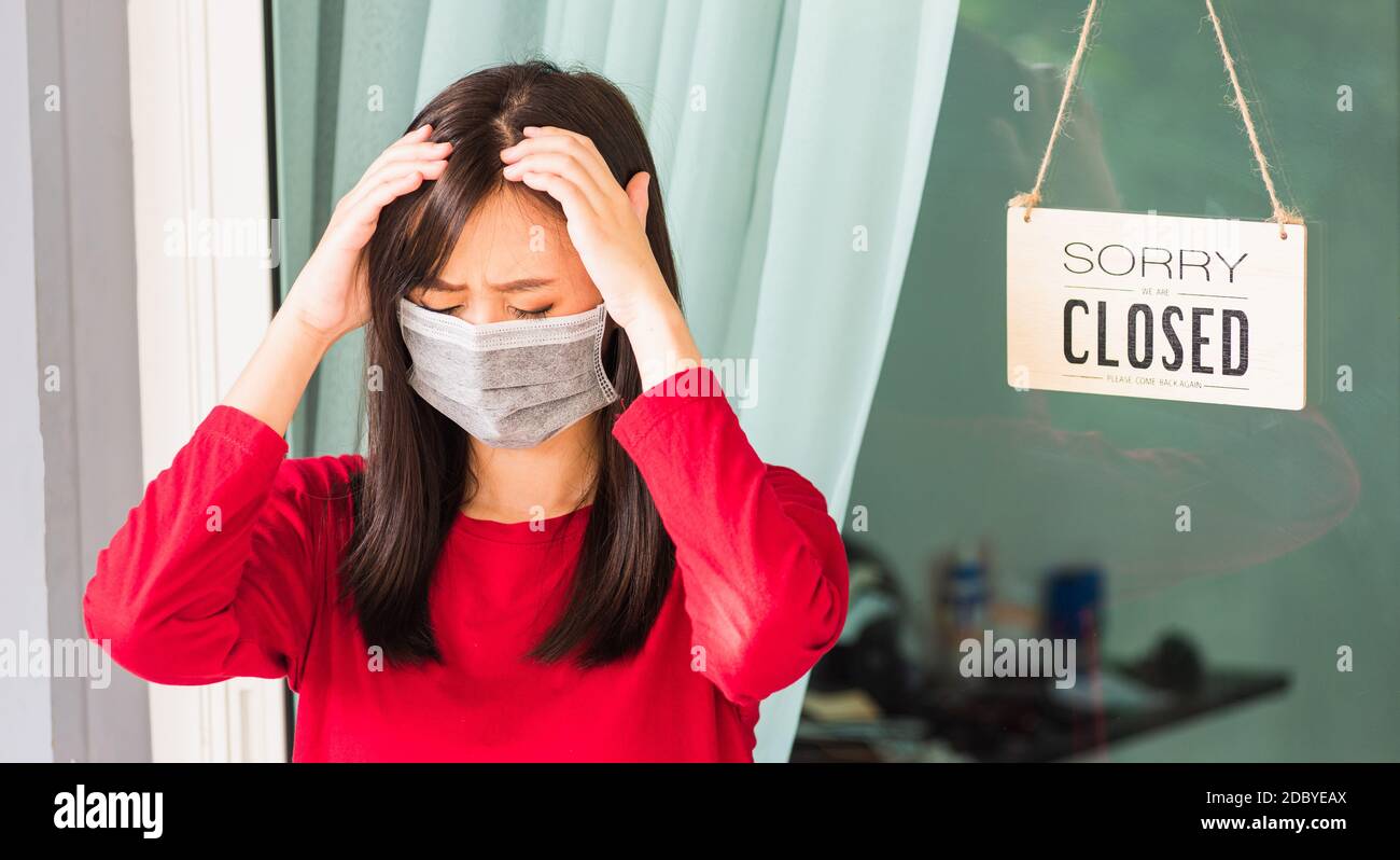 Asian young woman wear face mask protect she sad notice sign wood board label "SORRY WE ARE CLOSED PLEASE COME BACK AGAIN" hanging through glass door Stock Photo