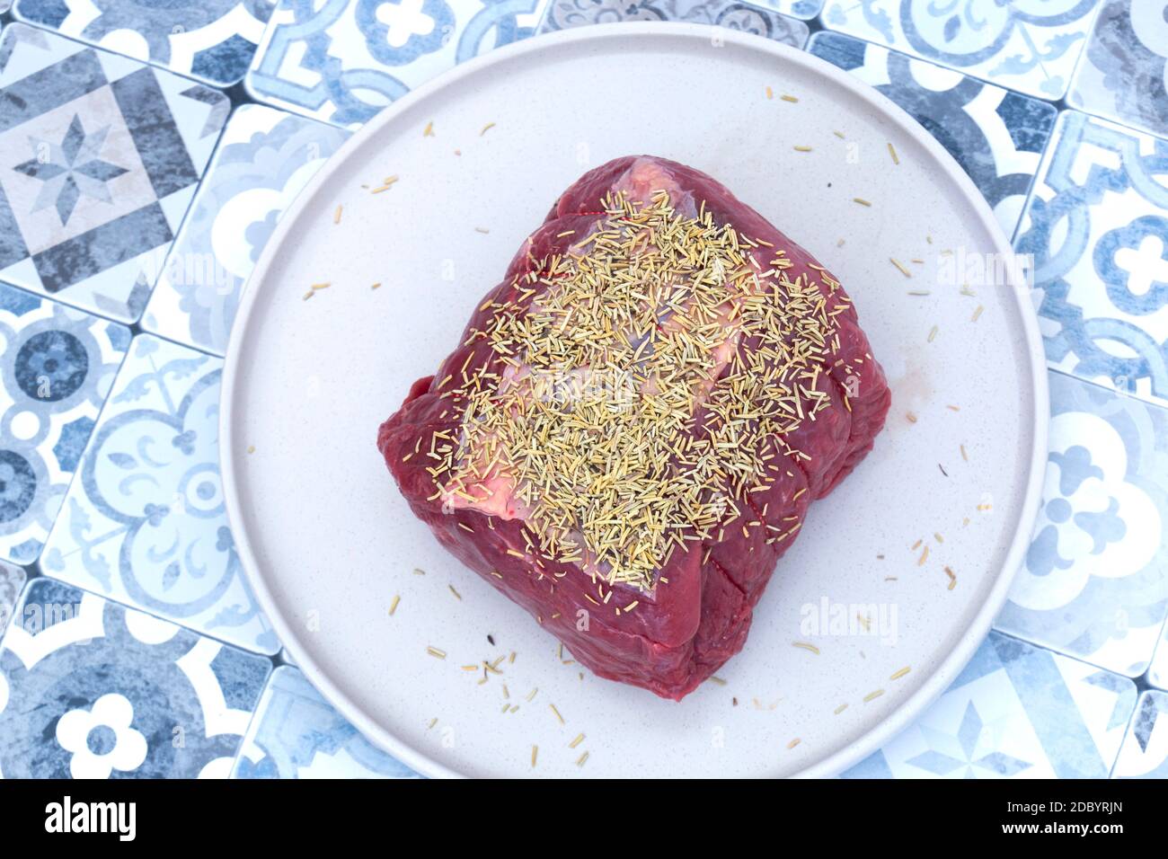 Raw beef roasting joint covered with rosemary on a grey plate. Blue