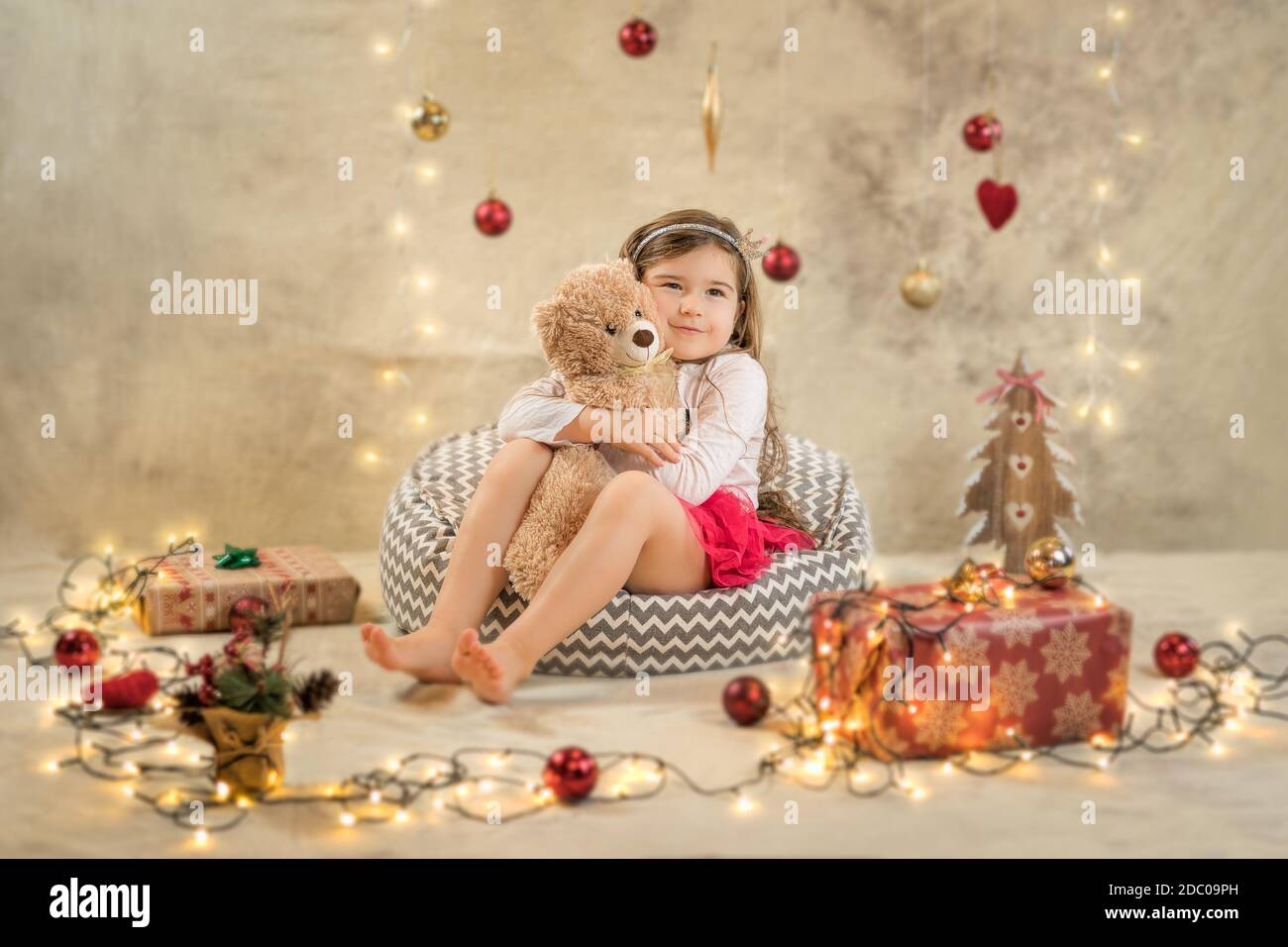 Christmas studio shoot of a cute baby girl that is hugging a teddy bear. Festive, beige background with gifts, Christmas balls and lights Stock Photo