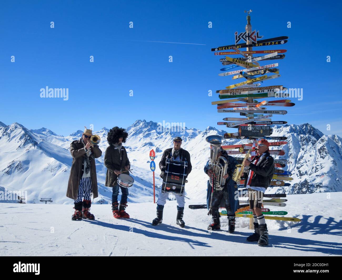 Music band in carnaval costumes playing at Ischgl-Samnaun ski resort with skis post pointing to international cities behind, Austria Stock Photo