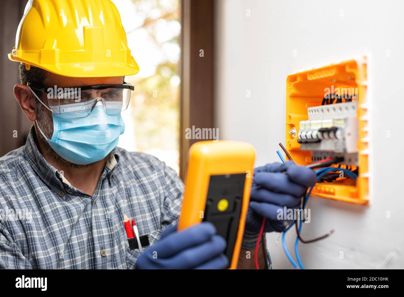 Electrician at work on an electrical panel protected by helmet, safety goggles and gloves; wear the surgical mask to prevent the spread of Coronavirus Stock Photo