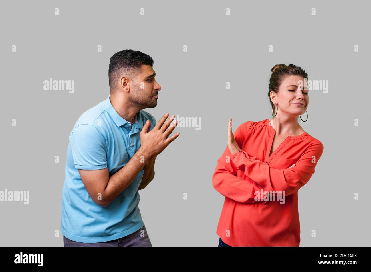 Portrait of funny couple in casual wear standing together, man holding arms in prayer and pleading forgiveness, looking with hope at naughty woman, is Stock Photo