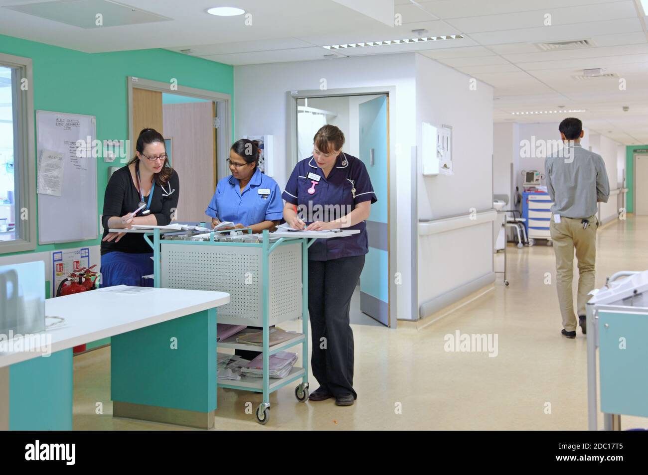 Modern UK hospital ward - nurses station. Two nurses discuss patient documents with a female doctor. Male doctor walking past. Stock Photo