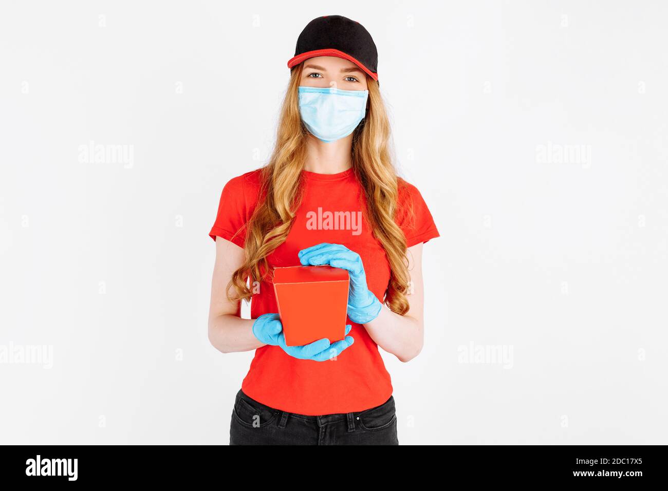 A uniformed courier wearing medical rubber gloves and a medical mask holds a paper box of food on a white background. Online stores, coronavirus, quar Stock Photo