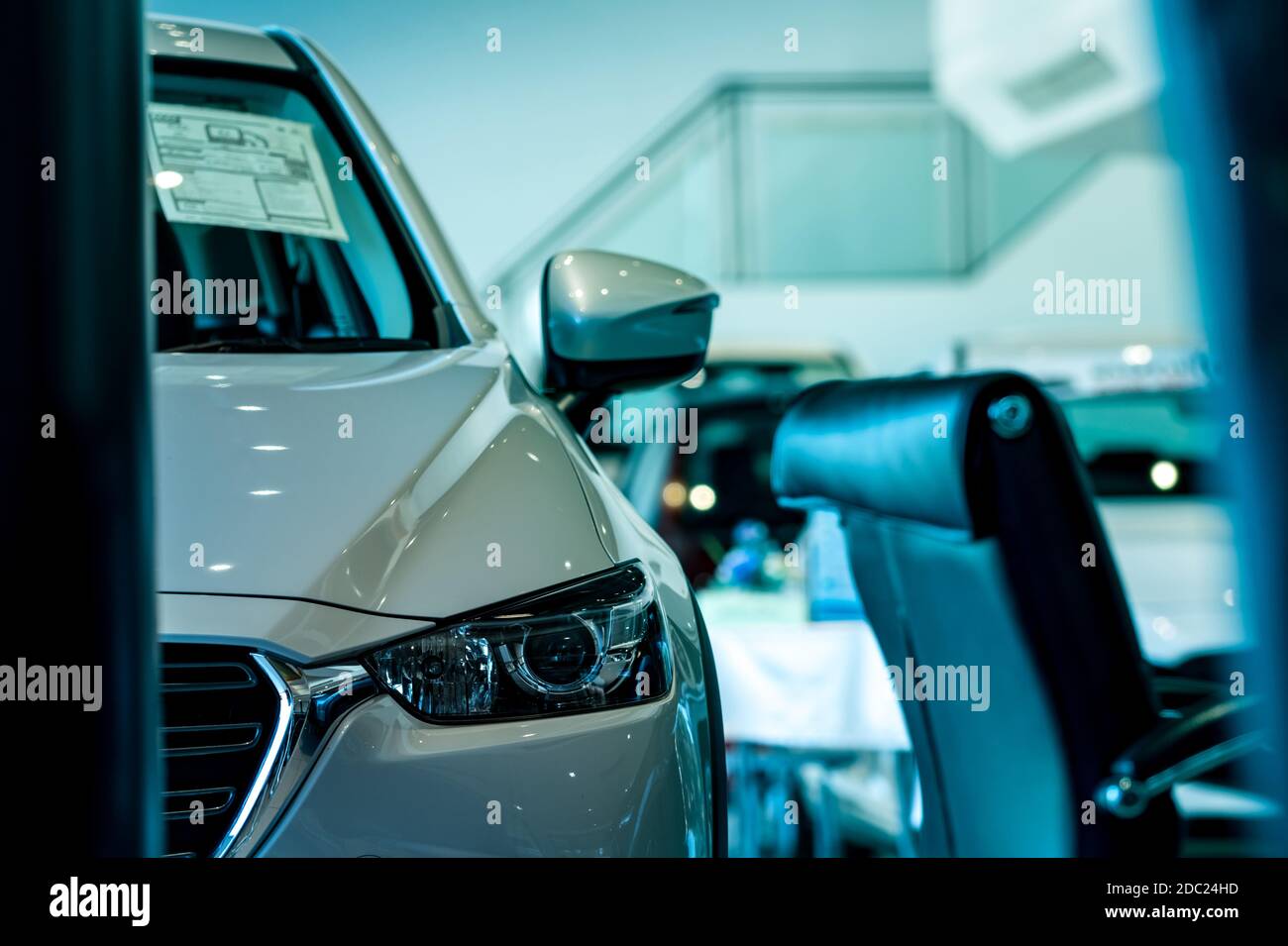Selective focus on headlamp of white shiny car parked in showroom. Front view of new car in modern showroom for sale. Car dealership concept. Electric vehicle business concept. Showroom interior. Stock Photo