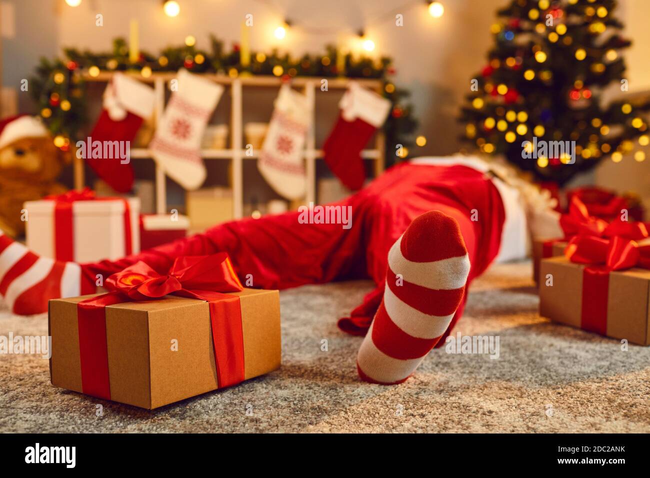 Gift box tied with red ribbon beside tired drunk Santa lying asleep on floor in living room Stock Photo