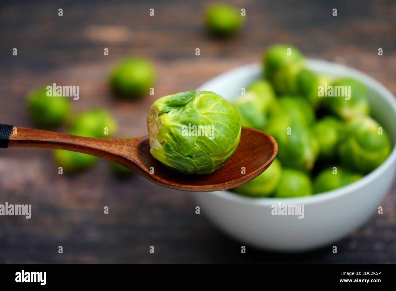 A wooden spoon holding a fresh Brussels sprout (German word: Rosenkohl). White bowl with Brussels sprouts in the blurred background. Stock Photo