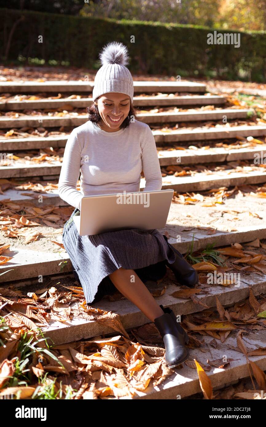 Woman sitting on an outdoor staircase. She is smiling using her laptop. Space for text. Stock Photo