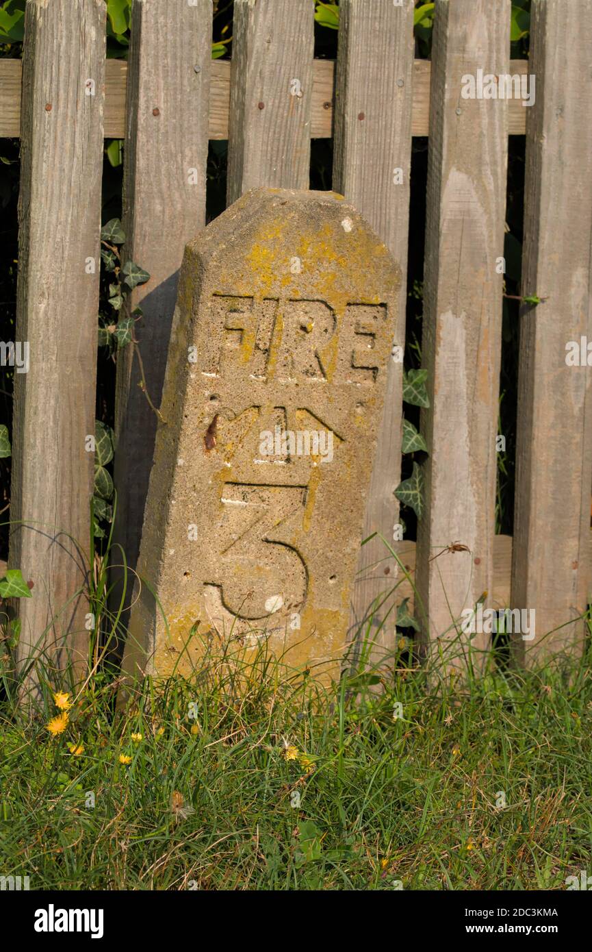 Unusual Concrete Fire Hydrant Marker In The Shape Of A Gravestone In The Style Of 1950's Marked With Fire And A Three In Front Of a Wooden Fence. Chri Stock Photo