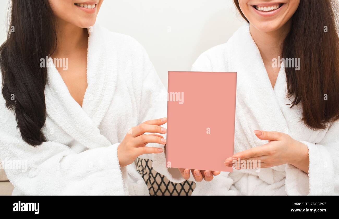 Two girlfriends wearing bathrobes holding an empty spa gift card for visits. Wellness spa advertisement Stock Photo
