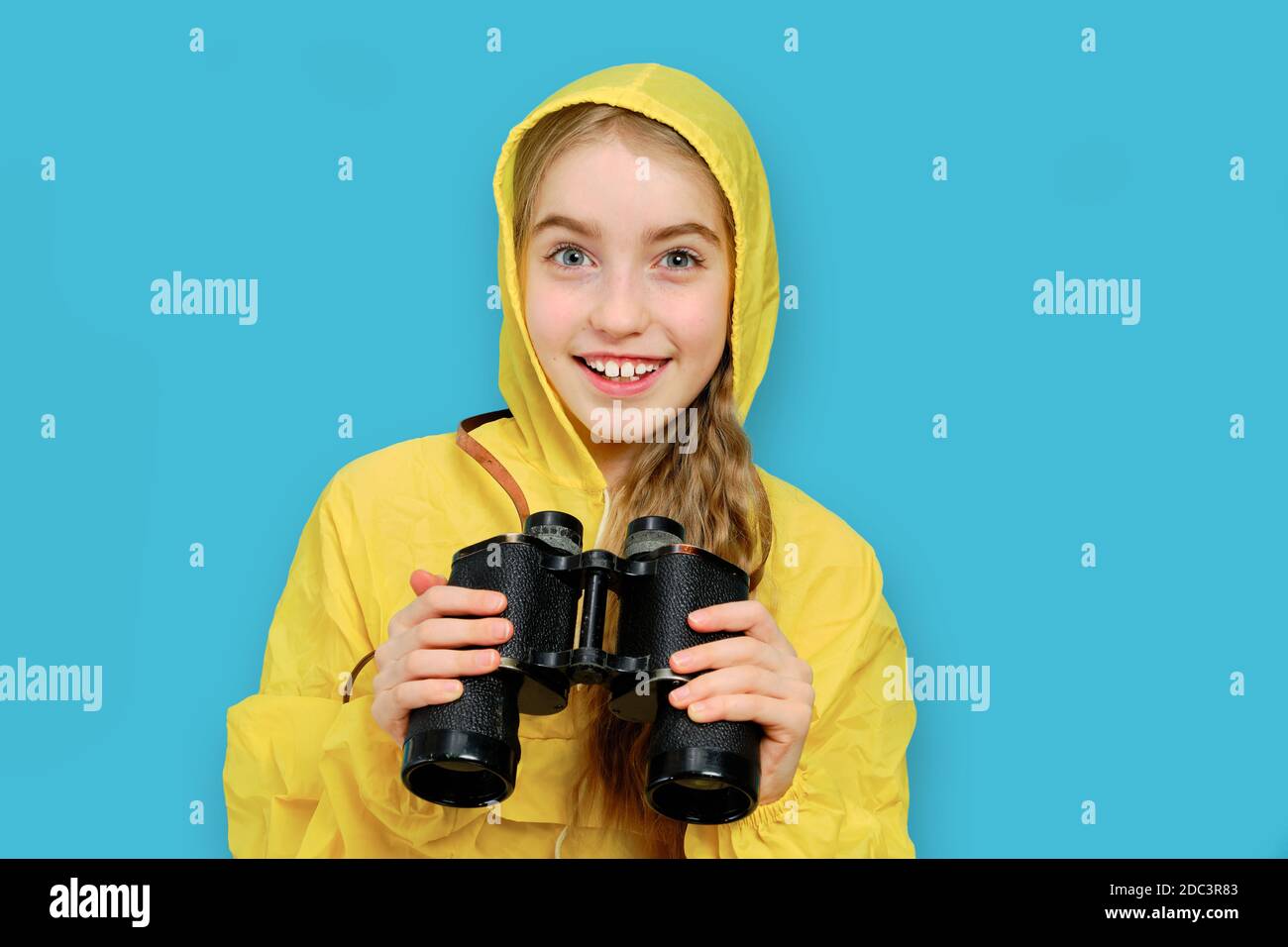 Caucasian joyful girl in a yellow jacket holds binoculars in her hands. hike idea and camping equipment.Studio isolated portrait on blue background. Stock Photo