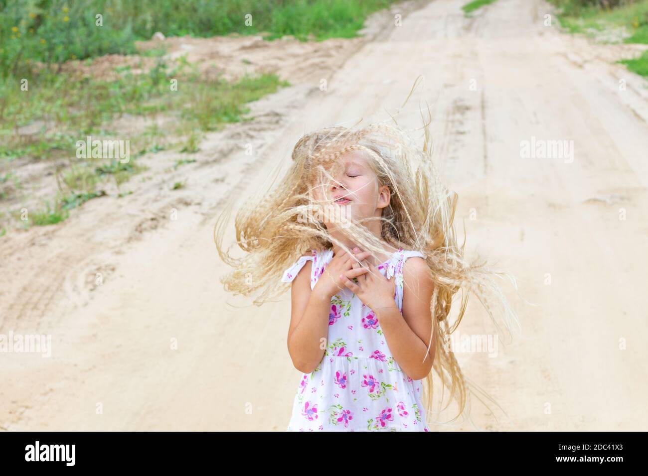 Beautiful girl with lush curly blond hair closed her eyes, folded her arms over her chest, flapped her hair, stands on sandy road. Feelings, emotions. Stock Photo