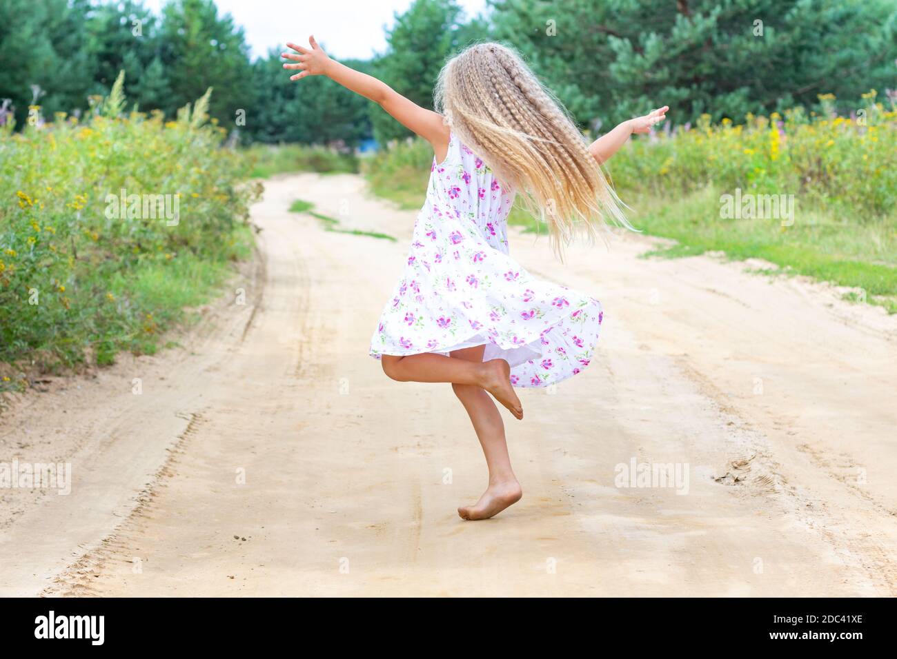 A girl with long blond curly hair is dancing, spinning on a forest road. Childhood, happiness, freedom of movement, dancing, reuniting with nature. Stock Photo