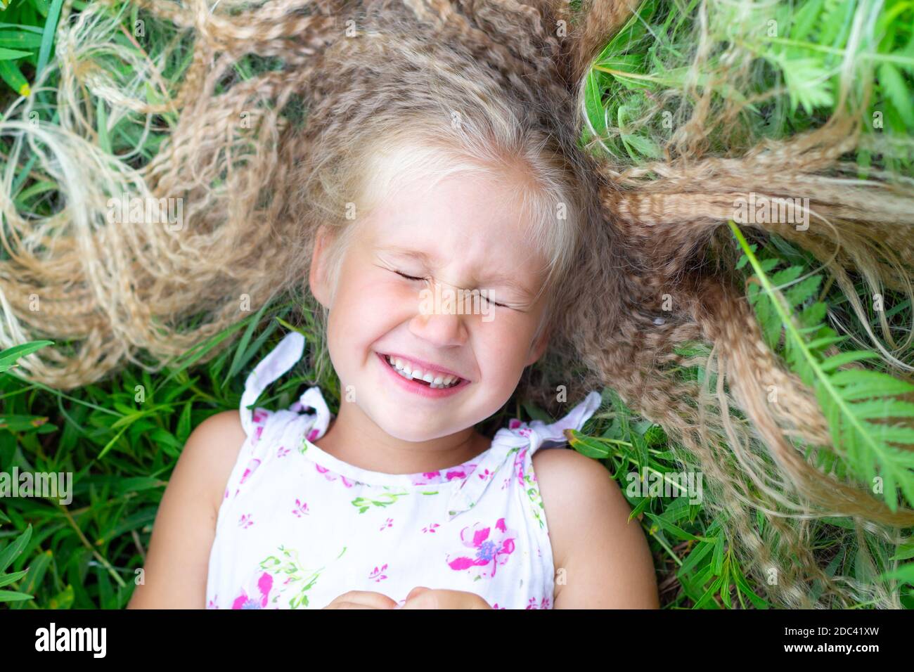 A little Caucasian girl with long blond hair in a white sundress lies on her back in the grass, closed her eyes, smiles, laughs. Childhood, happiness, Stock Photo