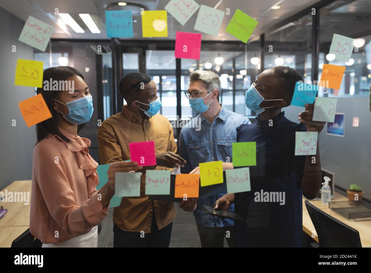 Diverse office colleagues wearing face masks discussing over memo notes on glass board at modern off Stock Photo