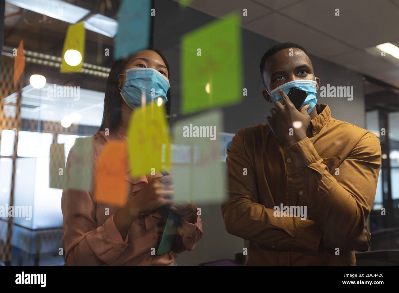 African american man and asian woman wearing face masks discussing over memo notes on glass board at Stock Photo