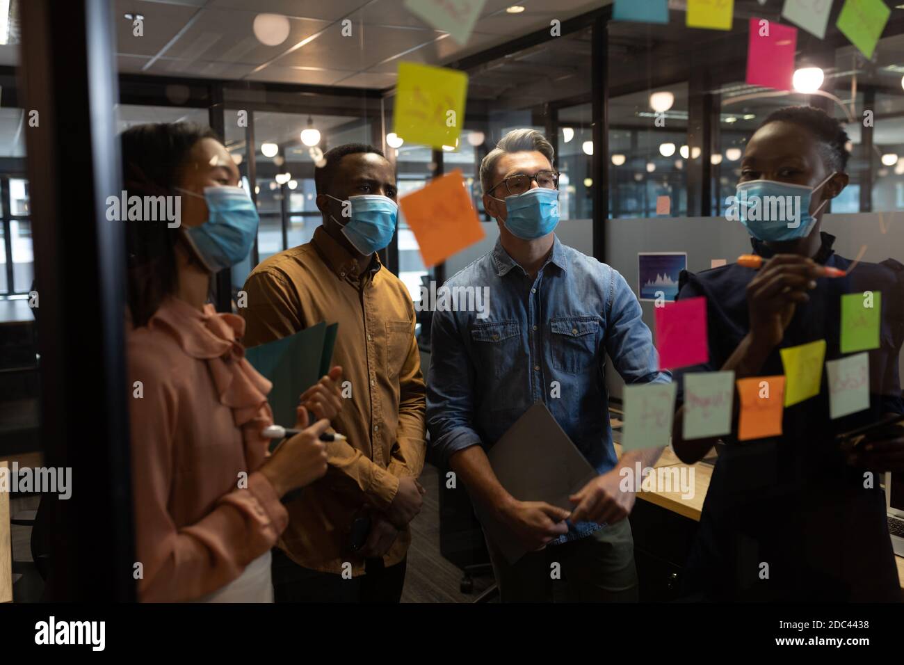Diverse office colleagues wearing face masks discussing over memo notes on glass board at modern off Stock Photo