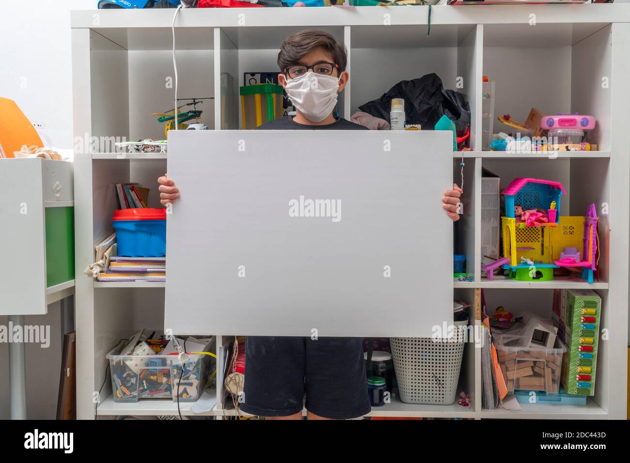 Covid-19 Stay at home concept: A little boy with glasses wears a face mask and holds a white empty board without a message. The kid stands Stock Photo