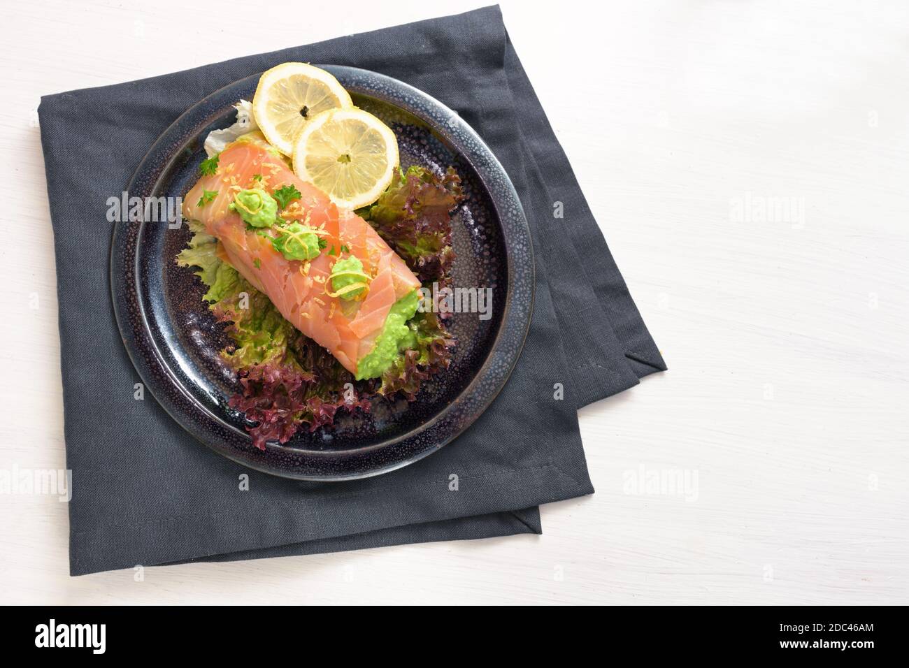 Smoked salmon filled with pea puree on lettuce salad with lemon slices and parsley garnish, dark plate and napkin on a white table, copy space, high a Stock Photo