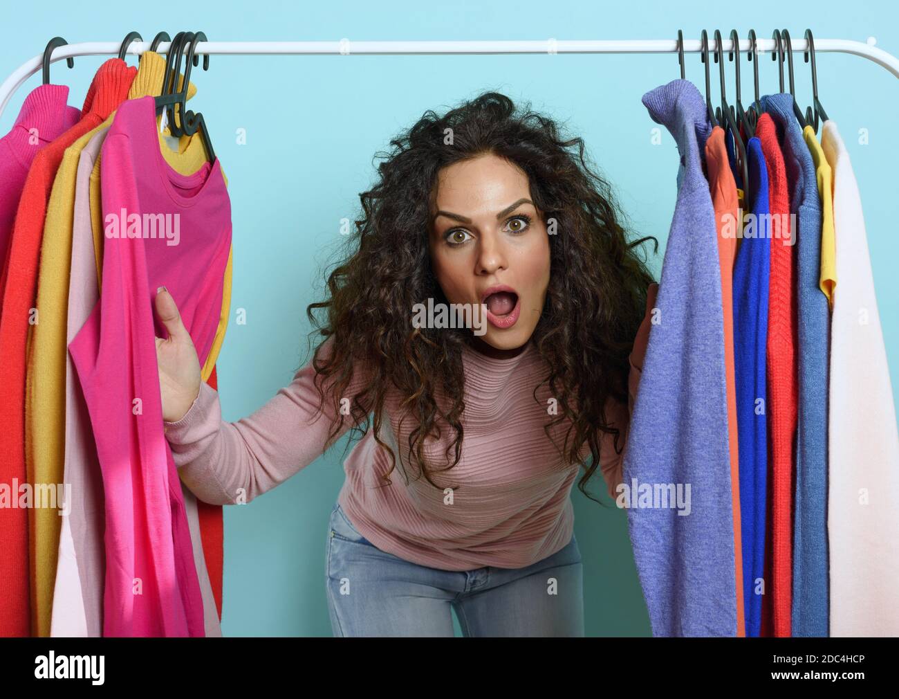 Woman with surprised expression chooses the clothes to buy in a store. Concept of shopping and shopaholic.Cyan background Stock Photo