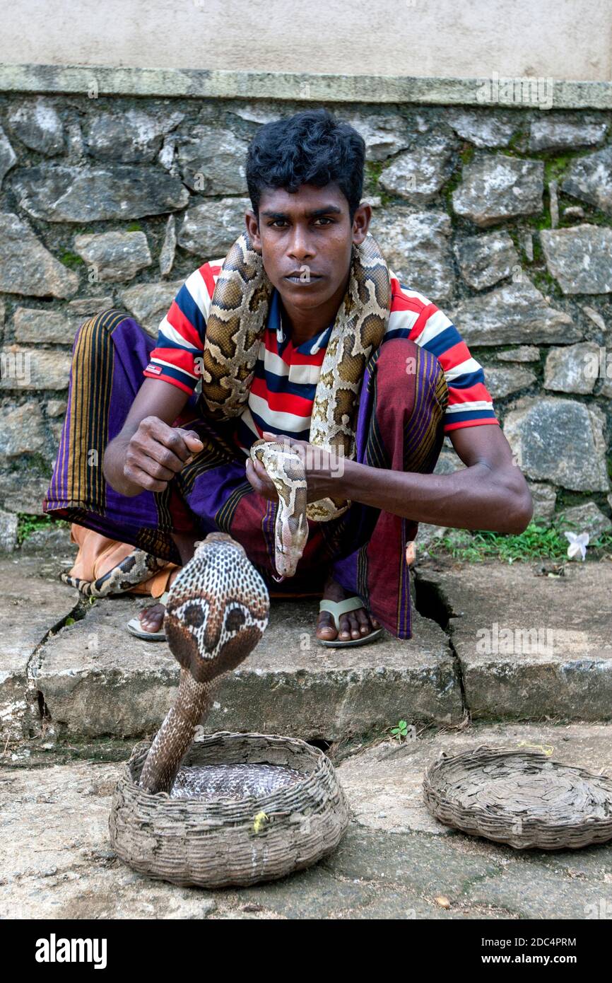 A young snake charmer with a python wrapped around his neck and a cobra in a cane basket sits on the footpath at Pinnawala in central Sri Lanka. Stock Photo