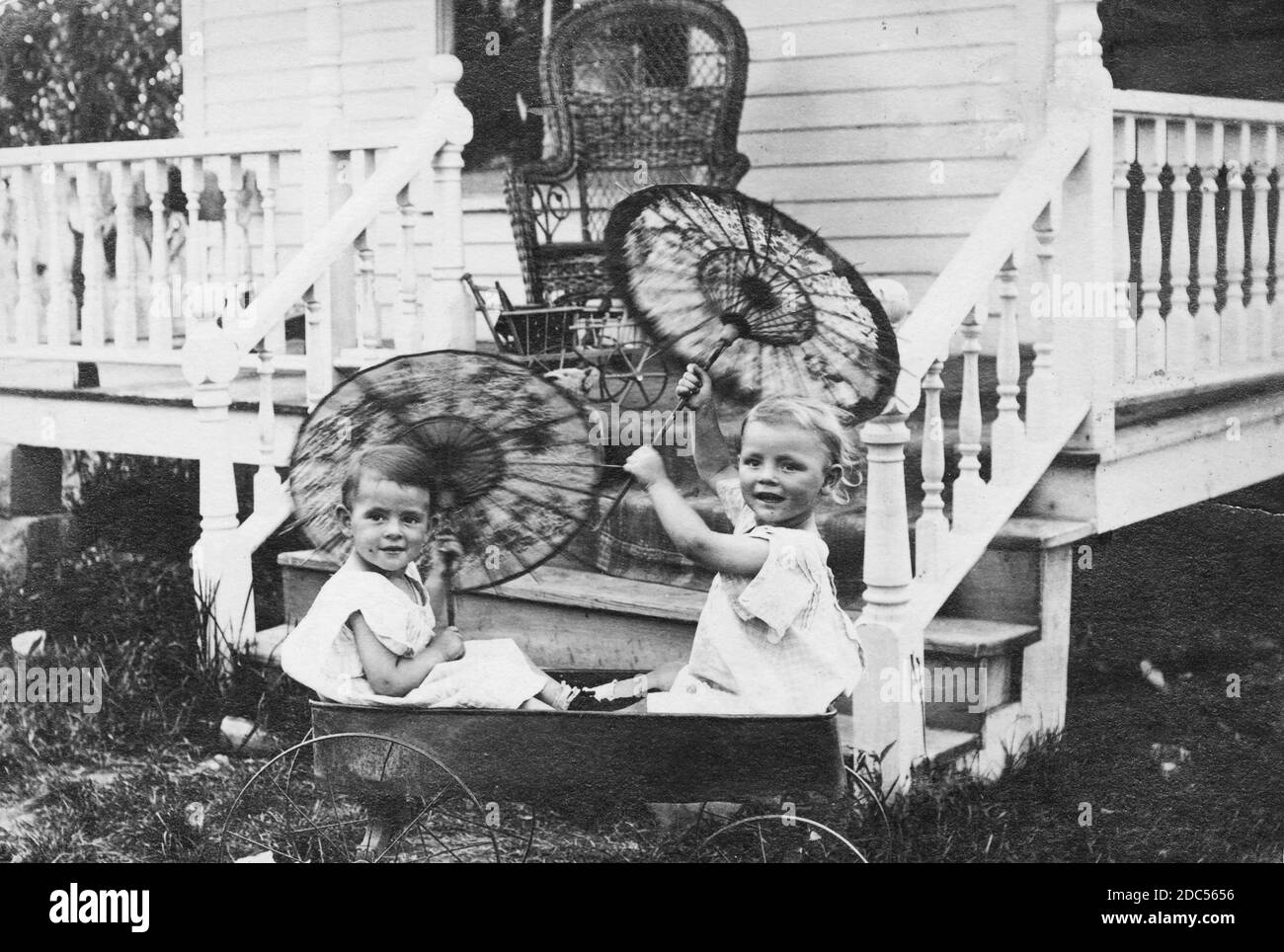 Two gleeful young children, a boy and a girl, twirl fancy parasols while sitting in a metal-wheeled coaster wagon beside their home's front porch, about 1906. Both kids are dressed in white dresses, which actually was normal wear for little boys of the early 1900s.  On the porch are a couple of other toys, partly obstructed, and an adult's wicker chair.   To see my kids-related vintage images, Search:  Prestor  vintage  kids Stock Photo