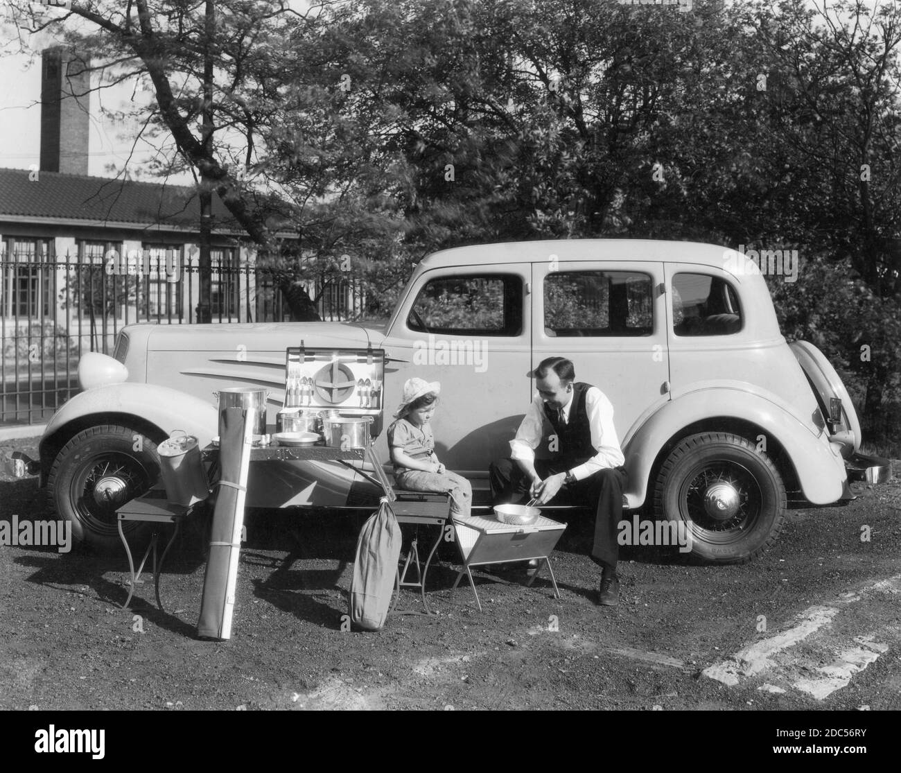 Camping gear and picnicking supplies, all neat and clean, are on display alongside a man and young girl in 1935. He sits on the driver's side running board of a shiny new Chevrolet 'Master Deluxe' sedan as she sits a type of folding chair. Visible are pots, pans, folding metal tables, a complete picnic set in a folding case, etc..   To see my transportation-related images, Search: Prestor  vintage vehicle Stock Photo