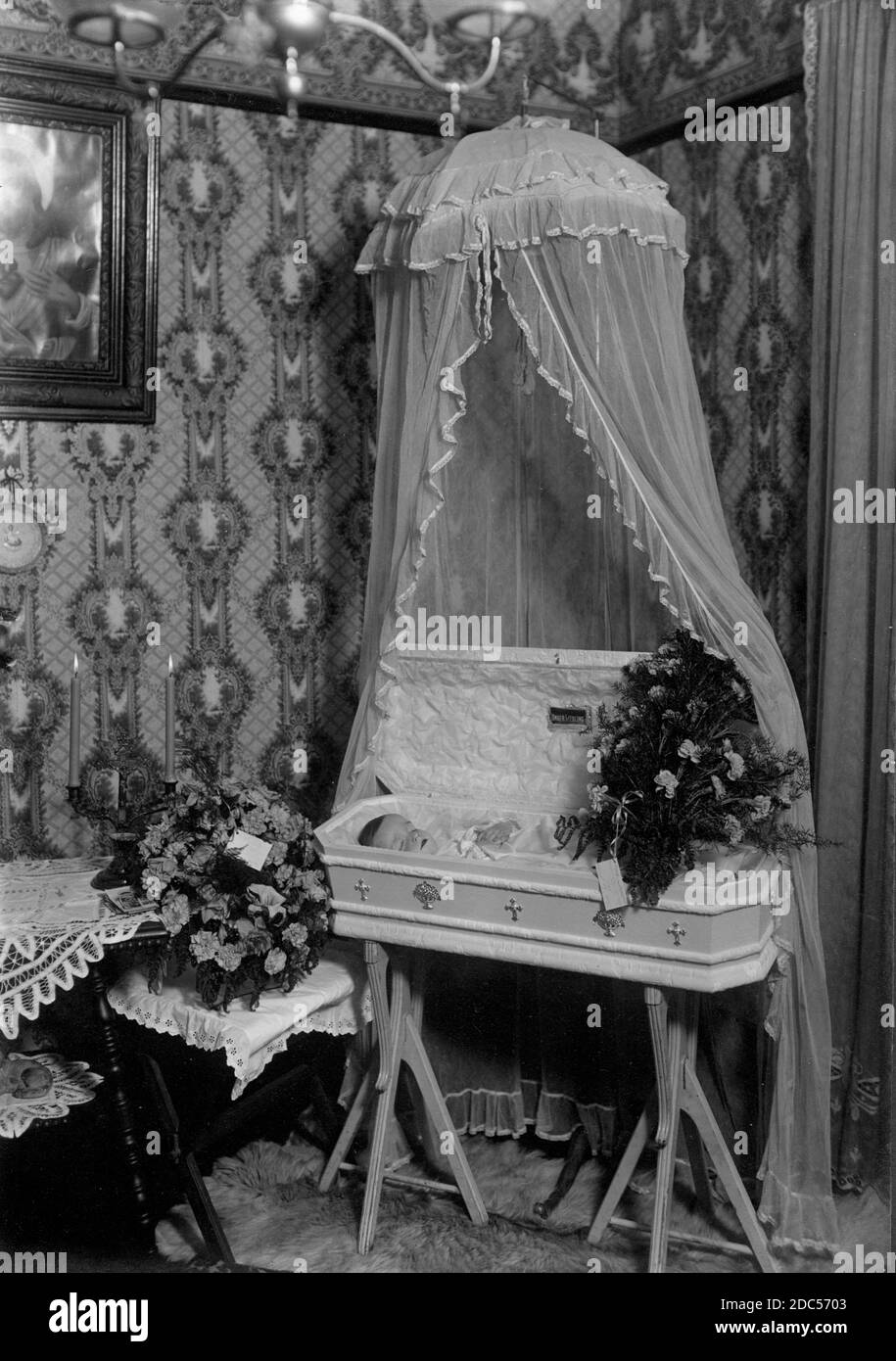 A small, deceased baby lies in a beautifully decorated, child-sized white coffin, about 1920. The coffin is displayed below a fancy white canopy suspended from the ceiling. This is all presented in the corner of the child's home, probably a main-floor parlor.  Inside the coffin's lid is attached a small plaque saying in German, 'Unser Liebling,' which means 'Our Darling.'  The casket sits on a pair of special tripod legs.   To see my kids-related vintage images, Search:  Prestor  vintage  kids Stock Photo