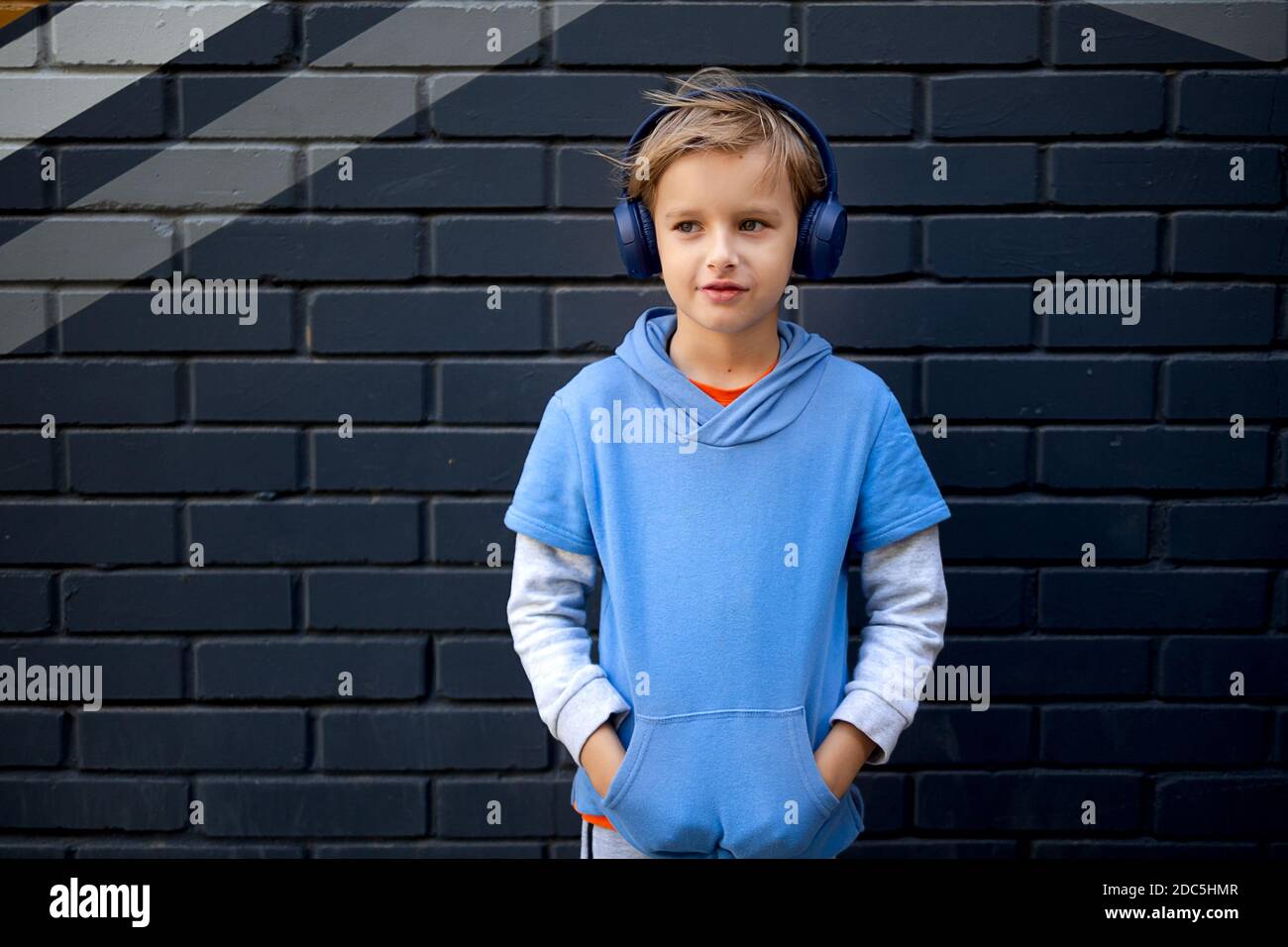 Blonde Caucasian boy in a blue hoodie and headphones stands on a city street against a gray brick wall, listening to music in a good mood Stock Photo