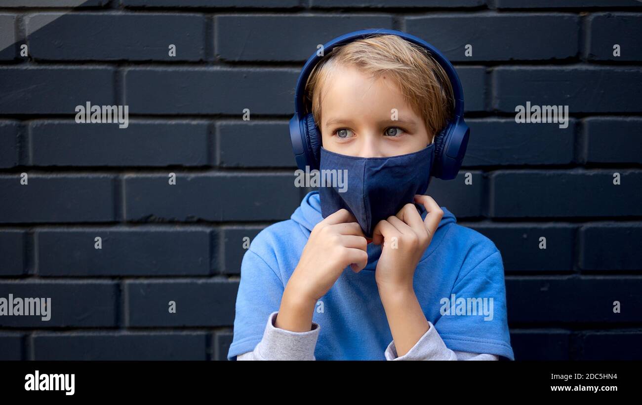 Portrait cute cool blonde caucasian little young boy in casual blue hoodie and medical protective mask stands city street against a gray brick wall Stock Photo