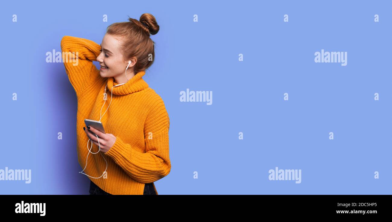 Adorable woman with red hair and freckles is listening to music on a blue studio wall with free space Stock Photo