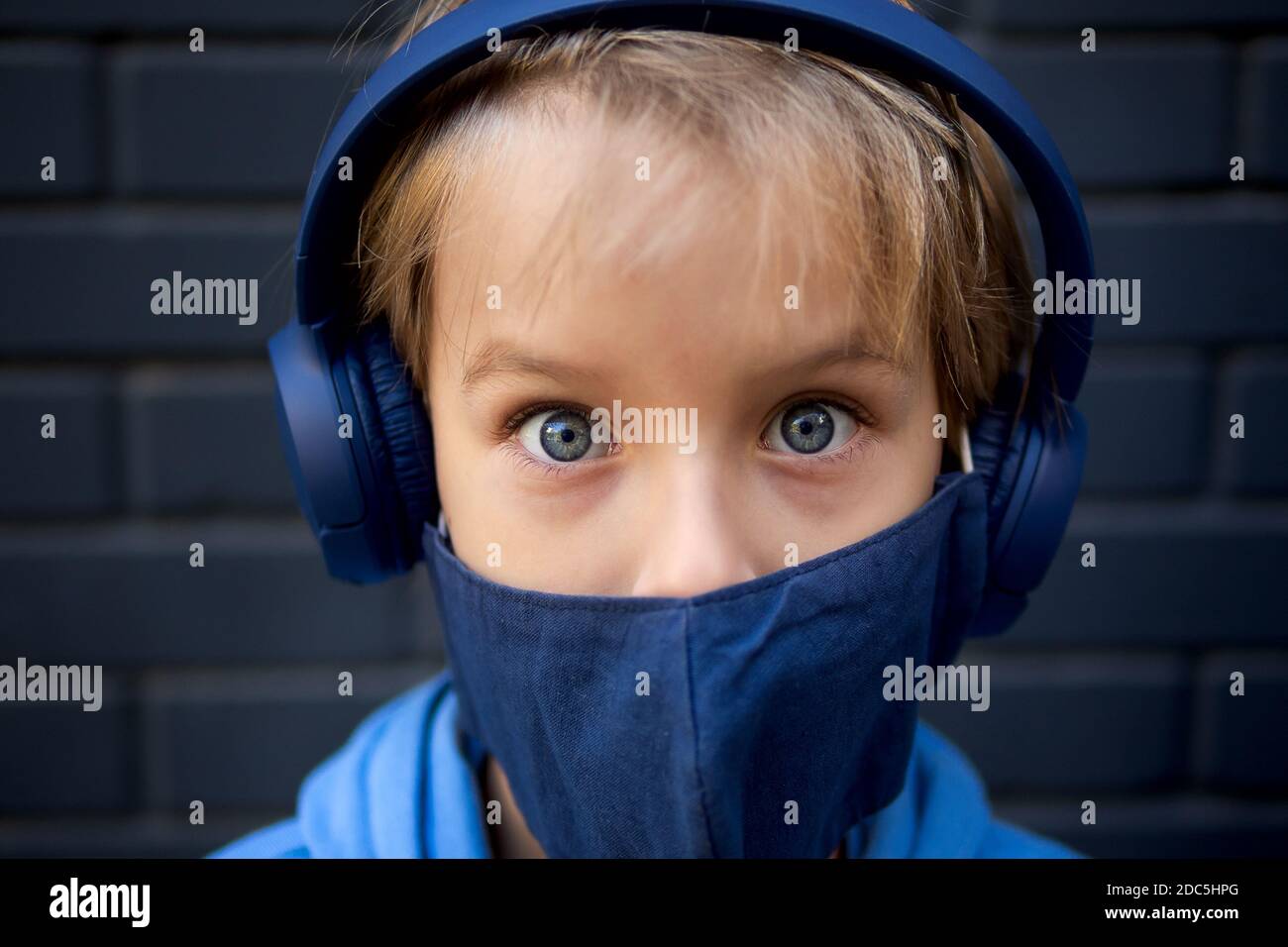 Close-up face portrait of a blonde caucasian boy. Looks at the camera. Dressed in a protective medical mask and blue headphones Stock Photo